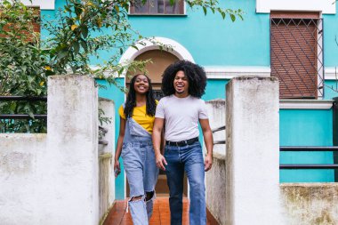 İki mutlu Latin arkadaş, şehvetli afro saçlı bir adam ve canlı mavi bir evin önünde yürüyen bir kadın..