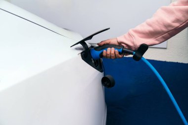 Tanımlanamayan birinin kolunun pembeyle yakın plan fotoğrafı. Beyaz elektrikli bir arabaya mavi bir elektrik şarj aleti takıyor..