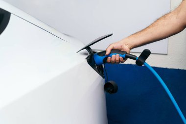 Mavi elektrik şarjını beyaz bir araca bağlayan tüylü kollu tanınmayan bir adam, çevre dostu teknolojiyi vurguluyor..