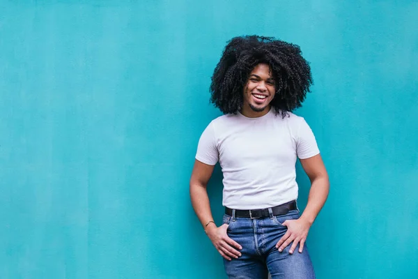 Afro-Latino man with curly hair laughing while standing casually in front of a turquoise wall, exuding happiness and style.