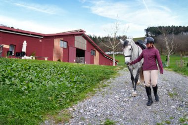 Atlı genç binici gri atıyla kırmızı bir ahır ve yeşil bir arazinin yanındaki yolda yürüyor..