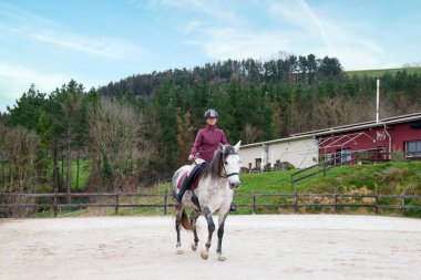 Miğferli ve binici kıyafetli genç sürücü kumlu bir arenada beyaz bir ata güvenle biniyor..