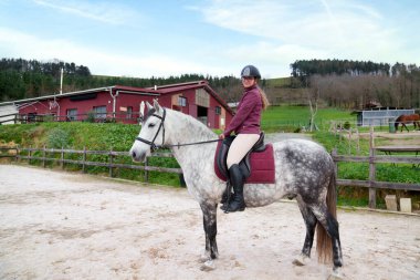 Benekli bir beyaz at üzerinde dengeli genç bir kadın, gezintiye hazır, manzaralı kırsal bir ahır arka planda.