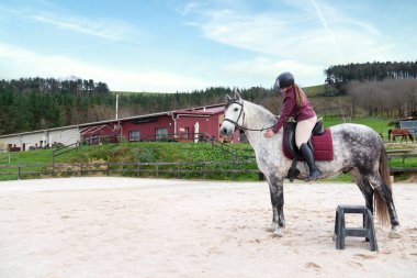 Kırsal kesimde, kumlu bir arenada benekli beyaz bir ata binerek pratik yapan genç bir binici..