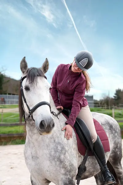 Güneşli bir günde güzel beyaz bir atı okşayan koruyucu kıyafetli neşeli genç bir sürücünün yakın çekimi..