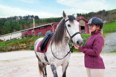 Dengeli genç bir binici beyaz atını ahırda tutuyor..