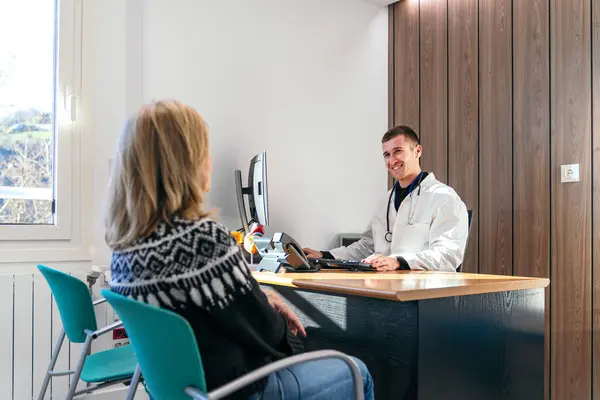 Olgun bir kadın modern bir klinikte masasında oturmuş, özenli erkek doktoruyla konuşuyor..
