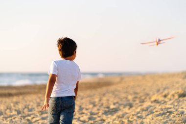 A child watches a toy airplane fly on a serene beach. clipart