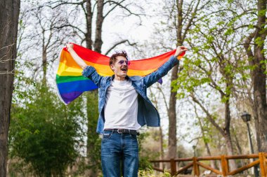 Büyük bir sevinçle, LGBT bayrağını yemyeşil bir park ortamında yükselten eşcinsel bir adam.
