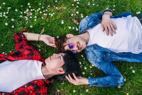 拉美和中国同性恋夫妇躺在一个鲜花繁茂的公园里 带着男女同性恋 双性恋和变性者的手镯示爱和自豪 免版税图库照片