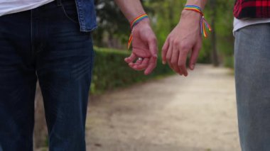 Sevgi dolu bir erkek çift tarafından tutulan renkli LGBT bileklikleri olan eller, sakin bir parkta duruyorlar..