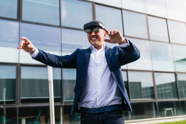 Takım elbise giyen ve sanal gerçeklik gözlüğü takan bir işadamı modern cam ofis binasının önünde sanal elementlerle etkileşime geçer..