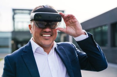 Takım elbiseli bir işadamı modern bir ofis binasının önünde gülümseyip sanal gerçeklik gözlüğü tutuyor..
