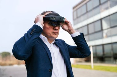 Takım elbiseli bir şirket lideri modern bir ofis binasının önünde sanal gerçeklik gözlüklerini ayarlıyor..