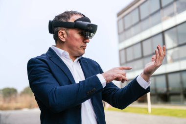 Takım elbiseli bir işadamı sanal gerçeklik gözlükleriyle modern bir ofis binasının önündeki sanal elementleri işaret ediyor..