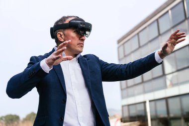 Modern bir ofis binasının önünde sanal gerçeklik gözlüğü kullanarak etkileşimli bir deneyim yaşayan takım elbiseli bir şirket lideri..