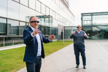 Sanal gerçeklik gözlüğü takan iki işadamı modern bir ofis binasının önünde sanal elementlerle etkileşime geçiyorlar..