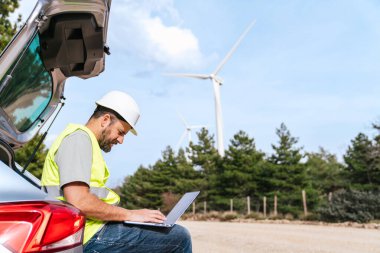 Yansıtıcı yelek ve şapka takan bir teknisyen arabanın bagajında oturur ve rüzgar türbinlerinin yanındaki dizüstü bilgisayarda çalışır..