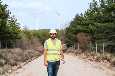 Rüzgâr türbini teknisyeni yüksek görüş açısına sahip yelek ve kasketli biri elinde dizüstü bilgisayarla kırsal bir yolda yürüyor. Arka planda rüzgar türbinleri var..