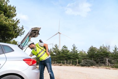 Yüksek görüşlü yelek ve miğfer giyen bir teknisyen, arkasında rüzgar türbinleri olan araçları hazırlamak için arabasının bagajını inceliyor..