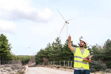 Yansıtıcı yelek ve başlık takmış bir teknisyen kırsal bir yolda rüzgar türbinini incelerken elinde bir pano ve el hareketi tutuyor..