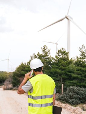Rüzgâr türbini teknisyeni telefonda elinde bir pano arka planda türbinlerle arka planda güvenlik yeleği ve kaskı olan bir adamla konuşuyor..