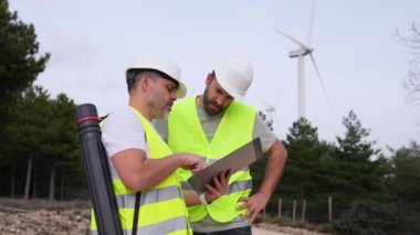 Yansıtıcı yelek ve kask giyen iki rüzgar teknisyeni türbinlerin yakınındaki bir pano ile bir projeyi tartışıyor..