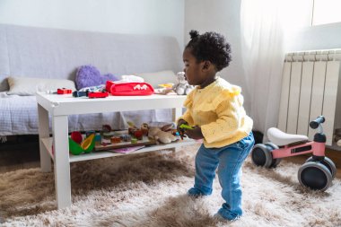 Genç bir kız masada oyuncaklarla oynuyor. Sarı bir kazak ve kot pantolon giyiyor.