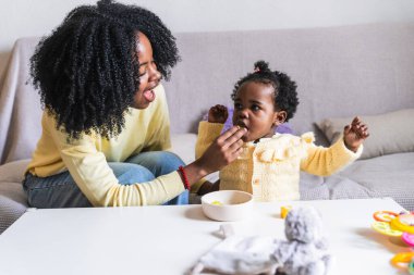Afrikalı bir anne bebeğini üzümle beslerken çocuk kanepede oyuncaklarla oynayıp neşeli ve besleyici bir an yaratıyor..
