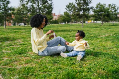 Bir kadın ve bir çocuk parkta çimlerin üzerinde oturuyorlar. Kadının elinde hava kabarcıkları var. Sahne huzurlu ve rahatlatıcı.