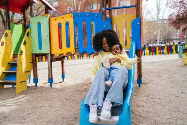 Mavi bir kaydırağın üzerinde bir kadın bir bebeği tutuyor. Kadın sarı bir gömlek giyiyor.