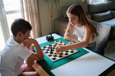 İki çocuk satranç oynuyor. Çocuklardan biri beyaz bir gömlek giyiyor. Diğer çocuk yerde oturuyor.