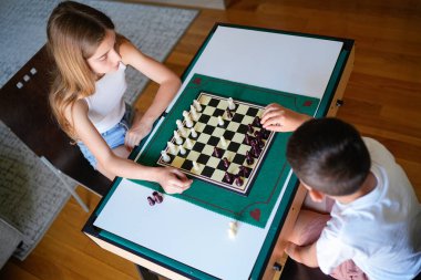 İki çocuk satranç oynuyor. Biri erkek, diğeri de bir kız. Yeşil tahtalı bir masada oturuyorlar.