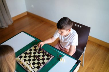 Bir erkek bir kızla satranç oynuyor. Çocuk beyaz bir gömlek giyiyor.