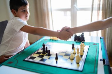 Bir çocuk satranç oynarken bir yetişkinle tokalaşıyor. Çocuk beyaz bir gömlek giyiyor ve yetişkin siyah bir gömlek giyiyor. Oyun tahtası yeşil ve siyah beyaz kareleri var.