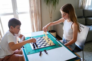 İki çocuk satranç oynuyor. Biri erkek, diğeri de bir kız. Yeşil tahtası ve parçaları olan bir masada oturuyorlar.