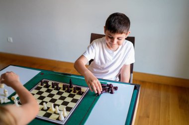 Bir çocuk başka bir çocukla satranç oynuyor. Beyaz gömlekli çocuk oynarken gülümsüyor.