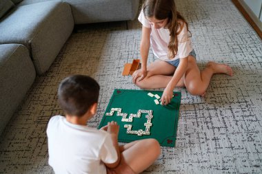 İki çocuk domino oynuyor. Biri yerde otururken, diğeri kanepede oturuyor.