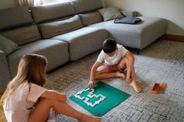 Yeşil bir tahtada domino oynayan iki çocuk. Çocuklardan biri beyaz gömlek ve şort giyiyor.