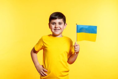 Genç bir çocuk Ukrayna bayrağı tutuyor ve gülümsüyor. Çocuk sarı bir arka planın önünde duruyor.