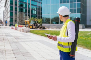 Yansıtıcı yelek ve başlık takmış kızıl saçlı bir kalite müfettişi inşaat işçilerini gözlemlerken not alıyor..