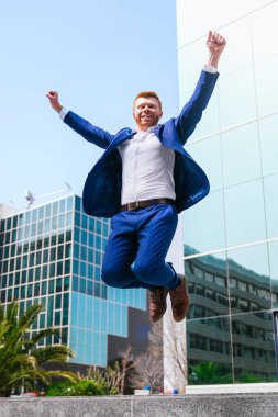 Takım elbiseli bir adam yüzünde kocaman bir gülümsemeyle havaya zıplıyor. Mutluluk ve heyecan kavramı, muhtemelen bir kutlama ya da zafer anını simgeliyor. Adamın kıyafeti, takım elbise de dahil.