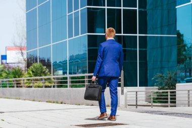 Mavi takım elbiseli genç, kızıl saçlı bir işadamının, iş parkında elinde siyah bir çantayla uzaklaşması. Profesyonellik ve iş anlayışı