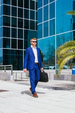 Genç, kızıl saçlı bir işadamı modern bir iş parkında elinde siyah bir çantayla güvenle yürüyor..