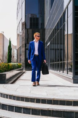 Genç, kızıl saçlı, güneş gözlüğü takan ve mavi takım elbiseli bir iş adamı elinde siyah bir çantayla bir iş parkında yürüyor. Profesyonellik ve resmiyet kavramı.