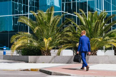 Mavi takım elbiseli genç bir profesyonel, bir iş parkında siyah bir evrak çantası taşıyan, modern cam bir binanın yanında yürüyor. Profesyonellik ve resmiyet kavramı.