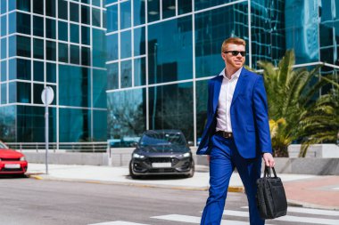 Mavi takım elbiseli kızıl saçlı bir işadamı, bir iş parkında evrak çantasıyla yürüyor. Güneş gözlüğü takıyor ve kendinden emin görünüyor..