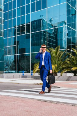 Mavi takım elbiseli kızıl saçlı işadamı, elinde çantayla caddede yürüyor, cep telefonuyla konuşuyor. Profesyonel ve odaklanmış görünüyor..