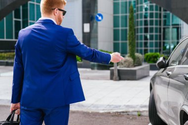 Mavi takım elbiseli kızıl saçlı bir işadamı elinde bavuluyla modern bir iş parkında siyah arabasının kilidini açıyor..