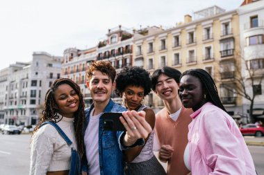 Çok ırklı bir grup insan cep telefonuyla selfie çekiyor. Hepsi gülümsüyor ve eğleniyor gibi görünüyorlar.